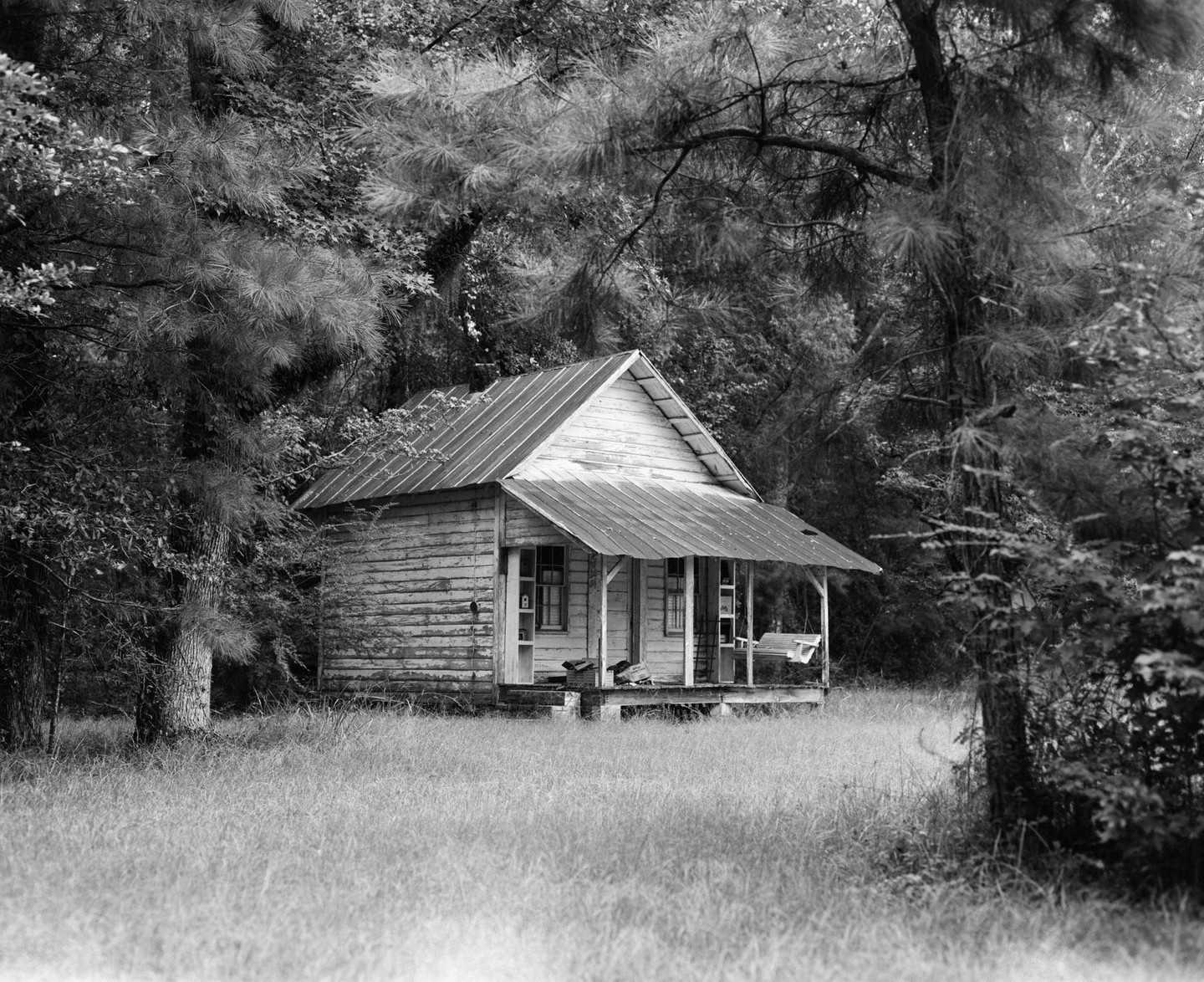 A small wooden house with a tin roof and porch stands in a grassy field surrounded by trees.