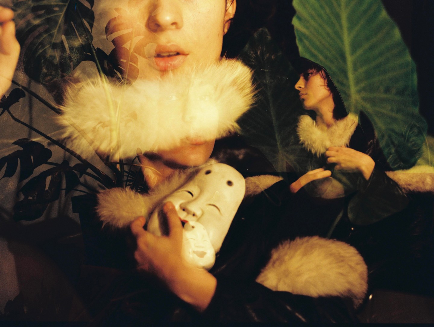 A woman in a fur-collared coat holds a mask, with a double exposure of her face and large leaves.