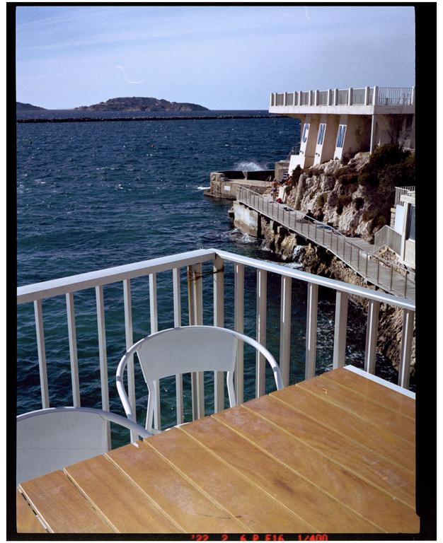 A wooden table and two chairs are on a balcony overlooking a rocky coastline with a building and the sea.