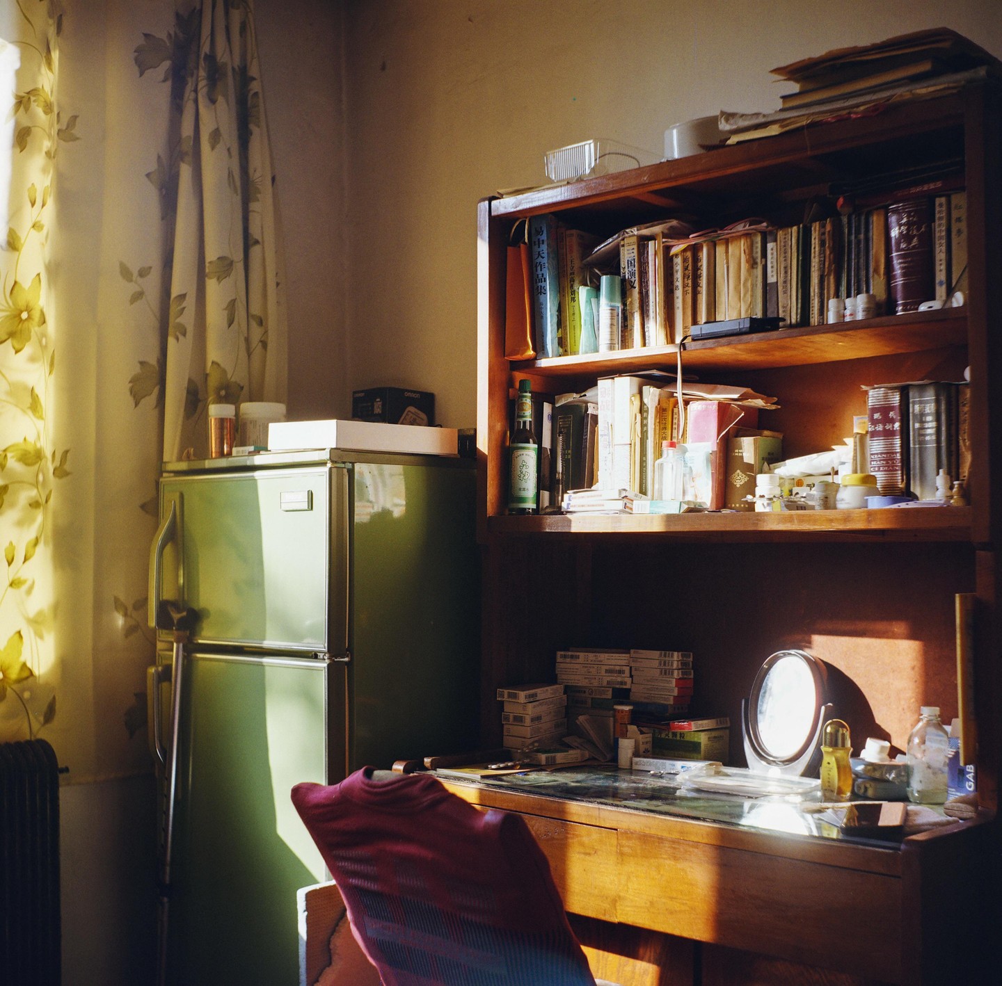 Sunlight illuminates a cluttered desk, bookshelf, and refrigerator in a room.
