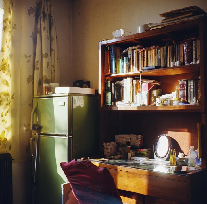 Sunlight illuminates a cluttered desk, bookshelf, and refrigerator in a room.