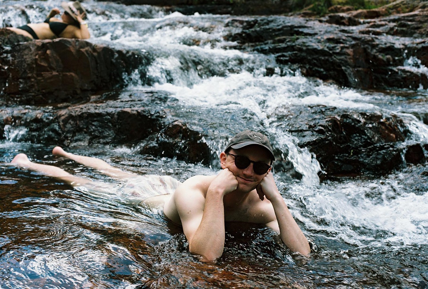 A man in a cap and sunglasses lies in a rocky river with a woman in a bikini in the background.