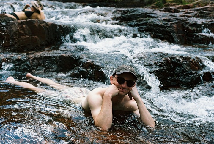 A man in a cap and sunglasses lies in a rocky river with a woman in a bikini in the background.