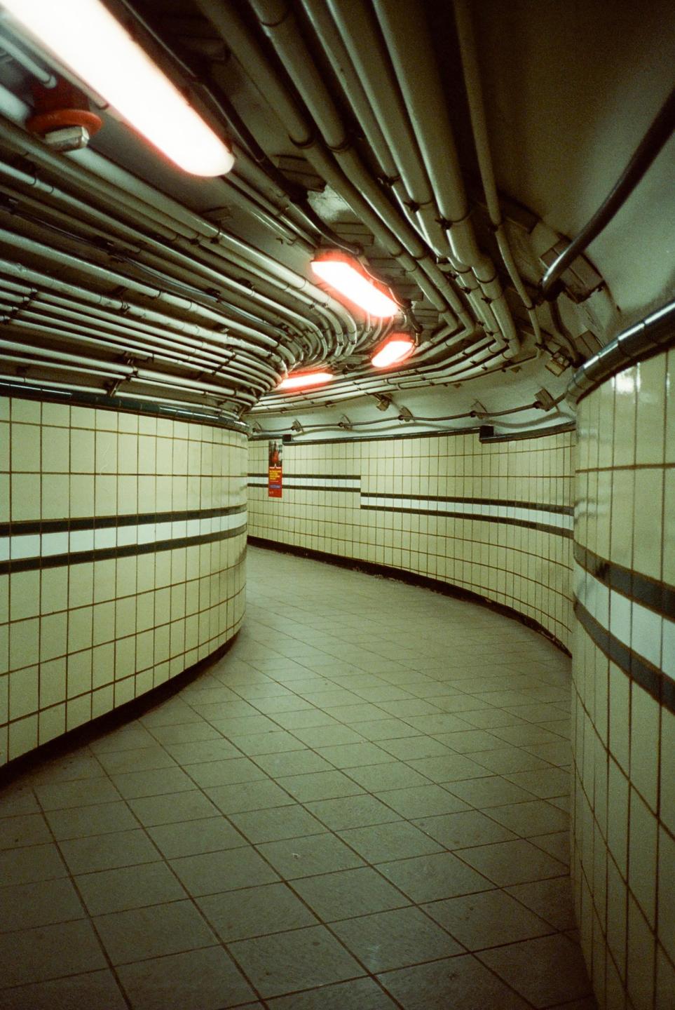 A curved underground tunnel with tiled walls and a ceiling covered in pipes and fluorescent lights is visible.