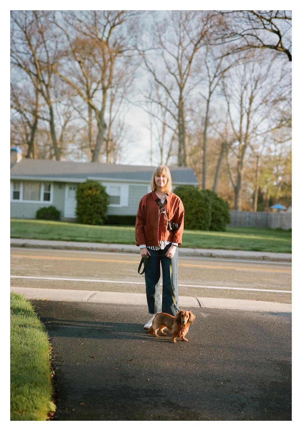 A woman stands on a paved surface holding a leash attached to a long-haired dachshund, with a house and trees in the background.