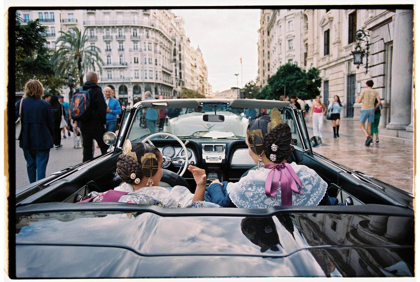 Two women in traditional dress sit in a convertible car on a city street.
