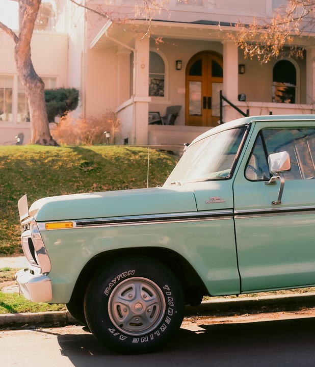 A light green pickup truck is parked on a street in front of a house with a wooden door and a porch.