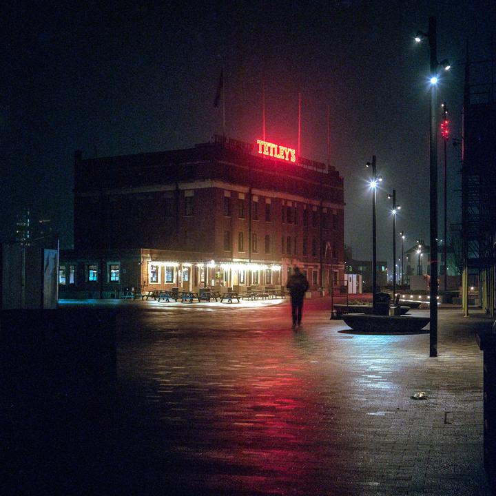 A person walks on a wet street at night with a building and neon sign in the background.