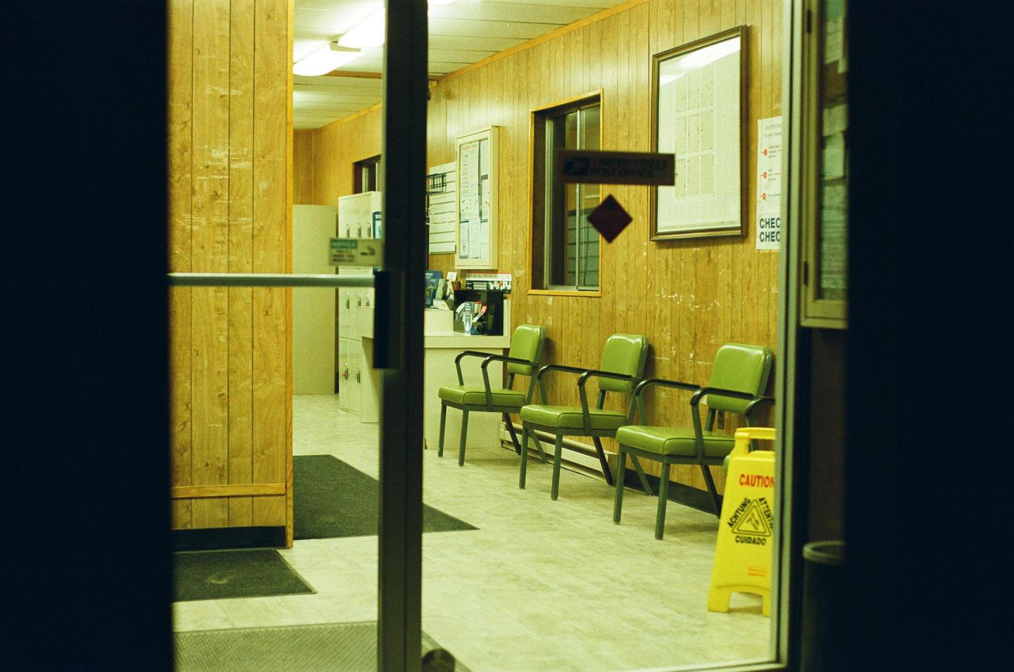 A waiting room with wood paneling and green chairs is viewed through a doorway.
