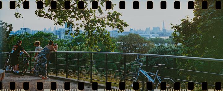 Three people stand by a railing overlooking a city skyline with a river and trees in the foreground.