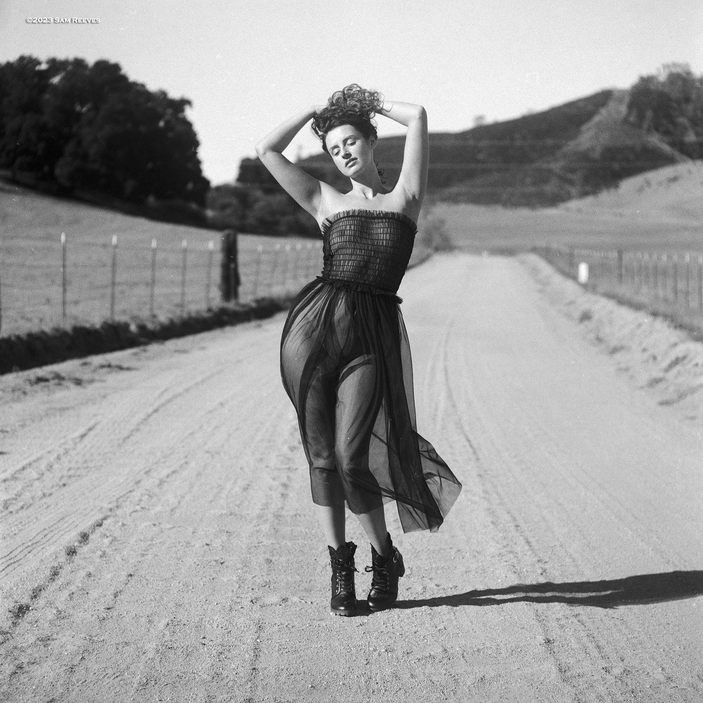 A woman in a sheer dress and boots stands on a dirt road with her hands behind her head.