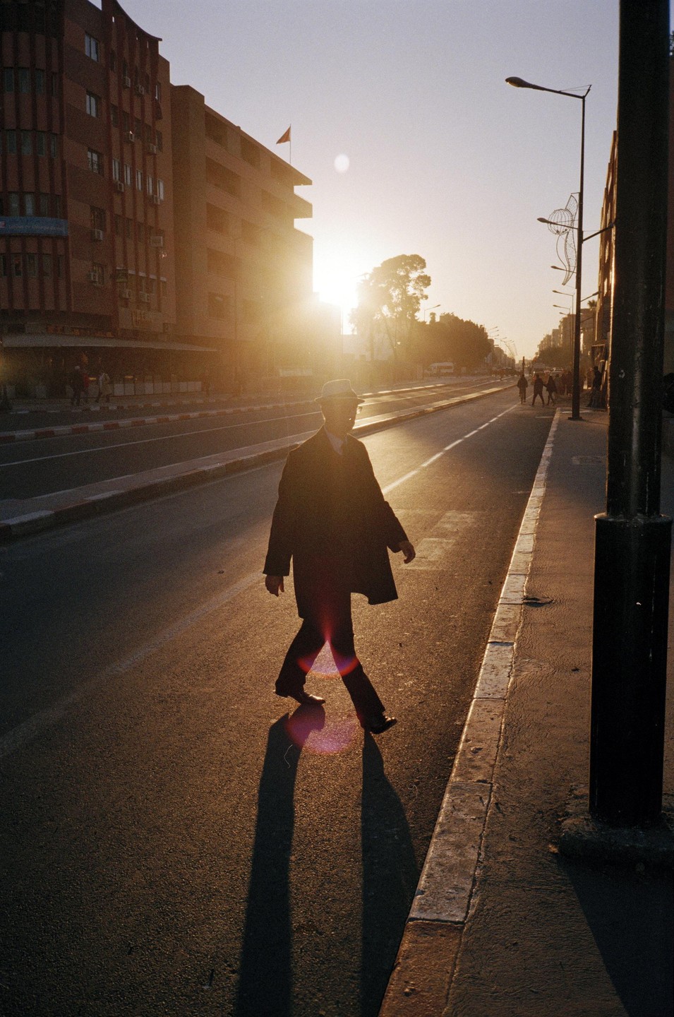 A man in a hat and coat walks across a street at sunset, casting a long shadow.