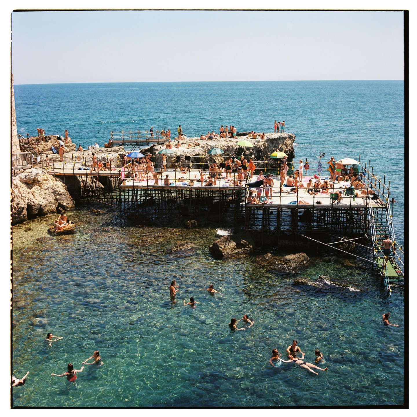 Many people are swimming and sunbathing on a rocky beach with clear blue water.