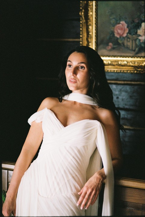 A woman in a white dress with a scarf around her neck looks up and to the right, with a painting in a gold frame behind her.
