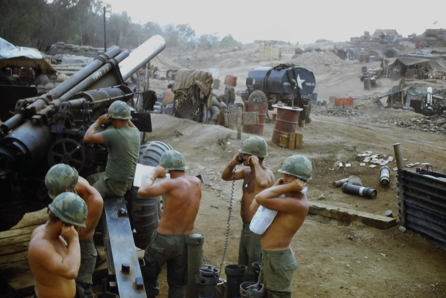 Several men in military fatigues and helmets are gathered around a large artillery gun in a dusty, open area with various military equipment and structures in the background.