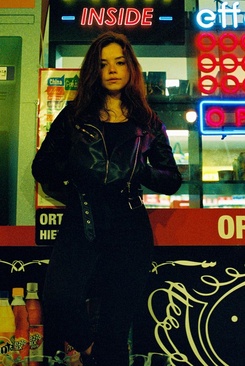 A woman in a leather jacket stands in front of a store window with neon signs.