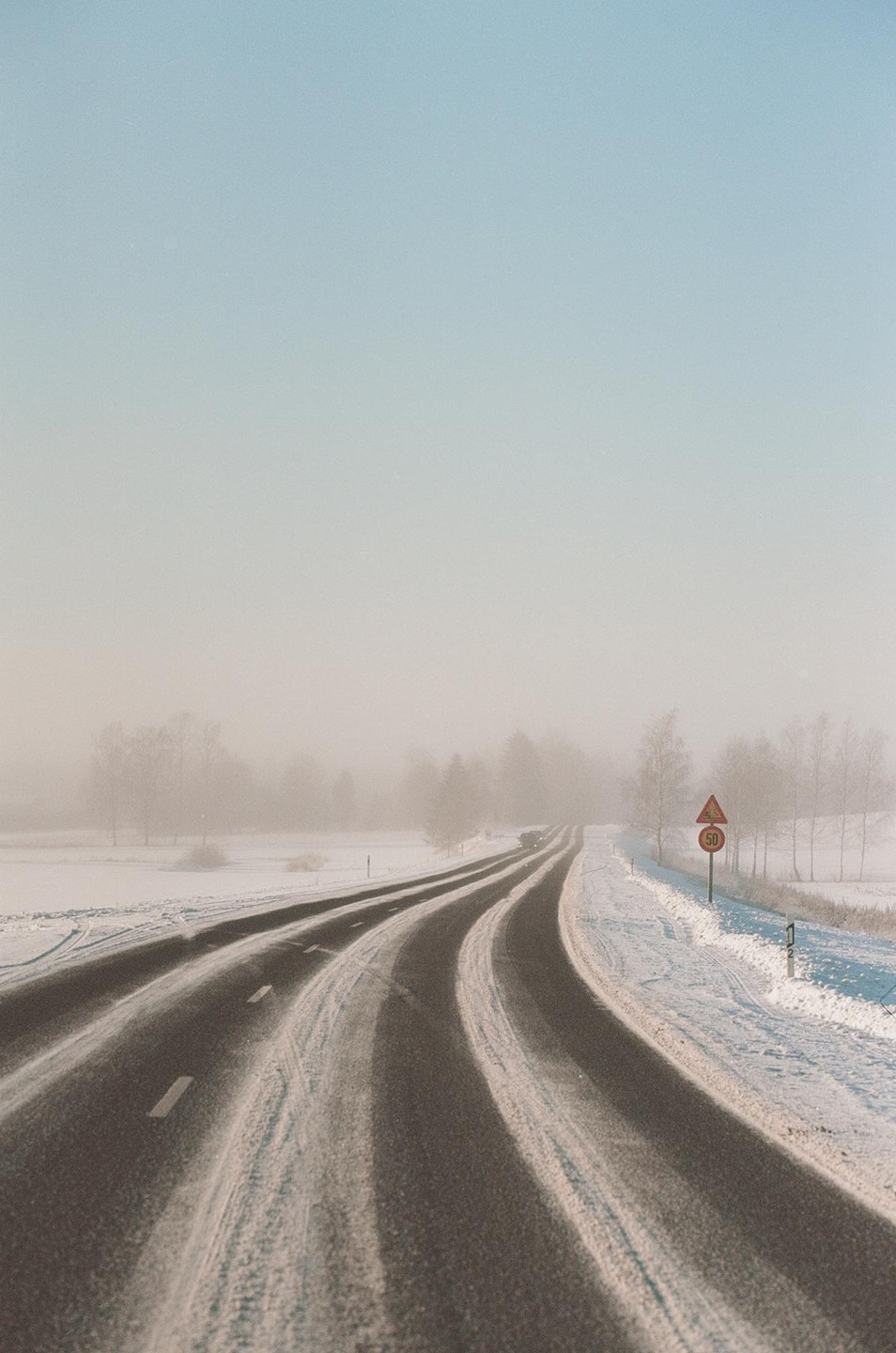 A snow-covered road curves through a hazy, winter landscape with trees in the distance.