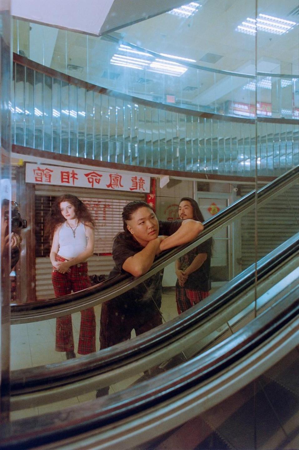 A man leans on an escalator railing while a woman and another man stand behind him, all reflected in a mirror.