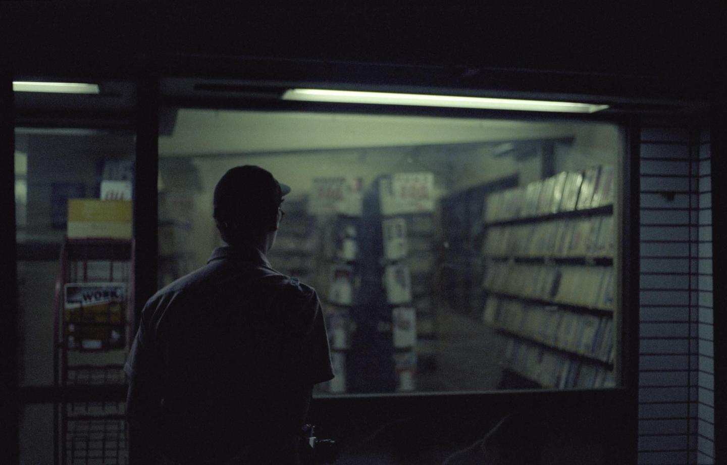 A man in a cap looks into a store at night, with shelves of items visible through the window.