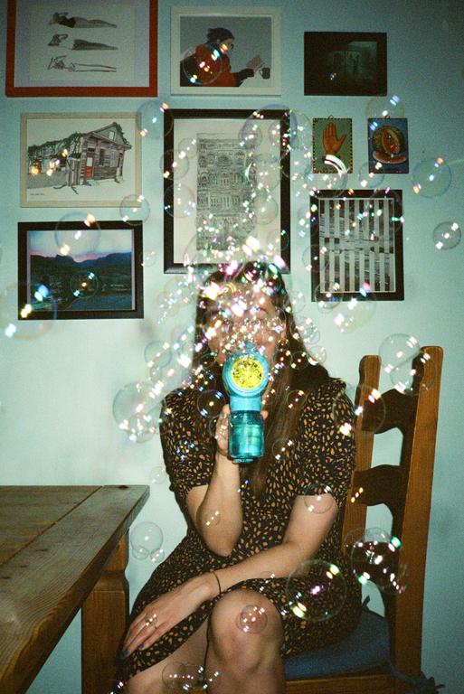 A woman sits at a table and blows bubbles from a toy bubble gun.