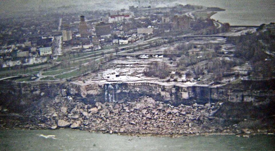 An aerial view shows Niagara Falls with a dry riverbed and a city in the background.