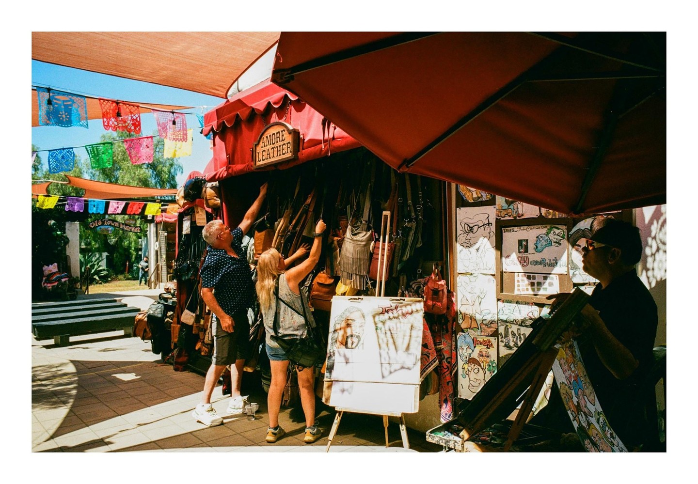 A man and a woman browse leather goods at a market stall under colorful banners.