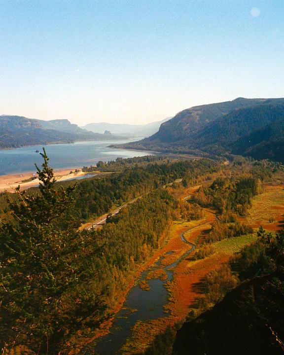 A winding river flows through a valley with autumn foliage, leading to a larger body of water surrounded by mountains.