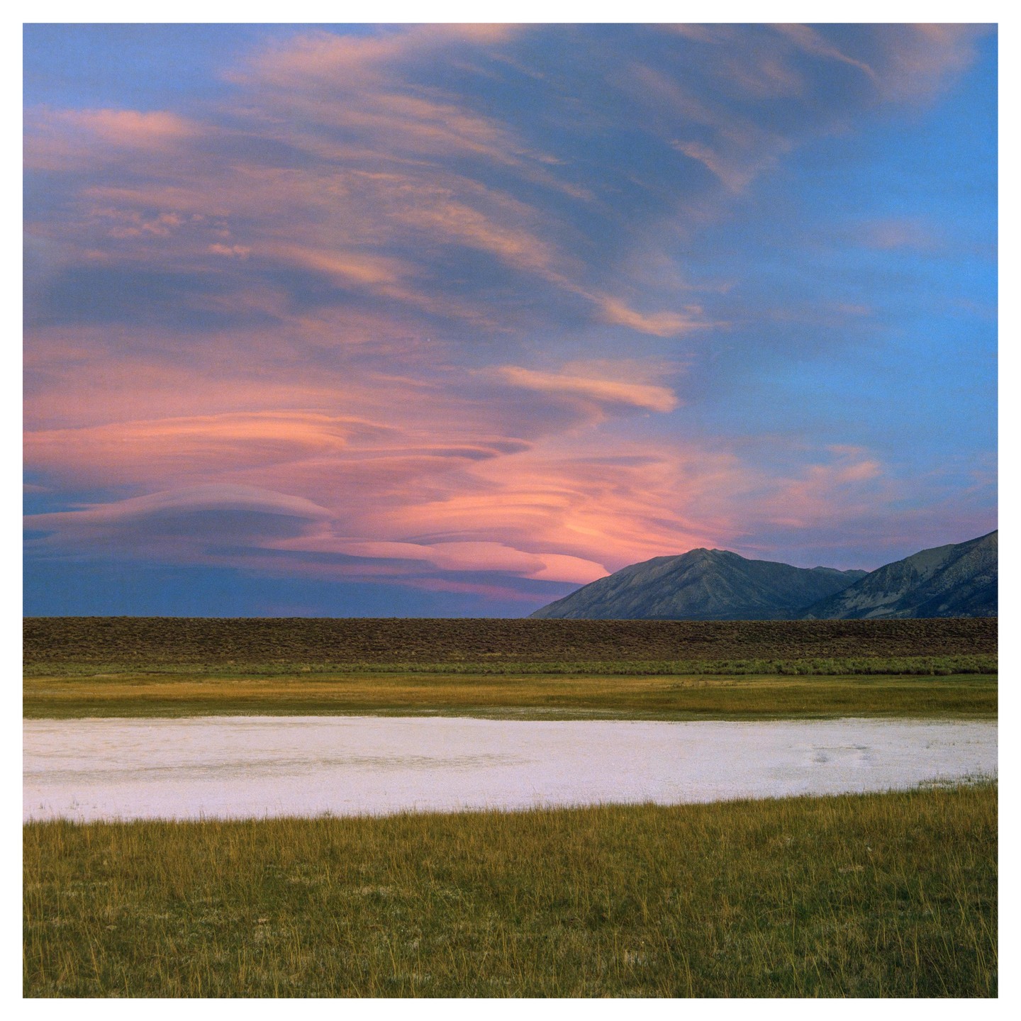 Pink clouds fill the sky above a mountain range and a field with a white pond.