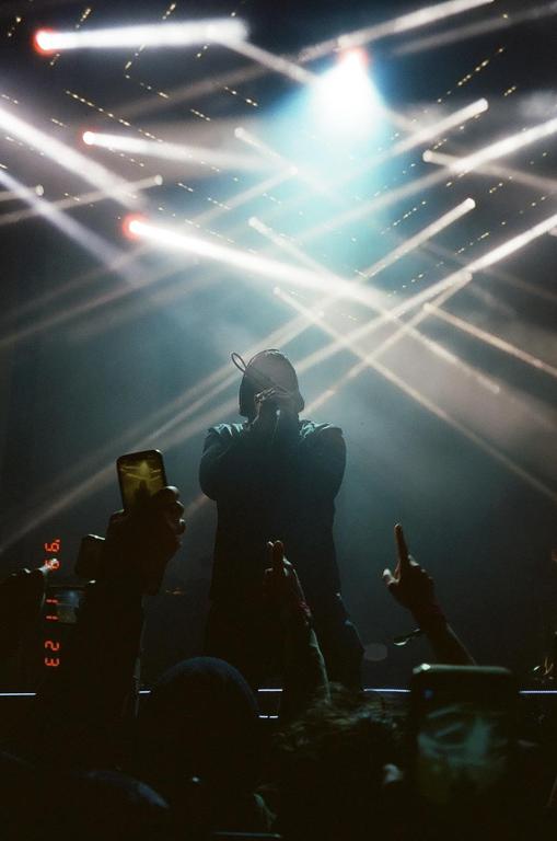 A musician performs on stage, illuminated by bright spotlights, as audience members hold up their hands and phones.