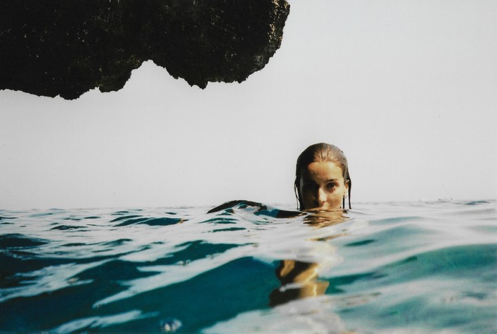 A woman with wet hair is in the ocean with a rock formation above her.