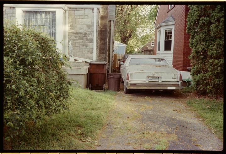 A white car is parked on a gravel driveway next to a house with a stone facade and a red brick house.