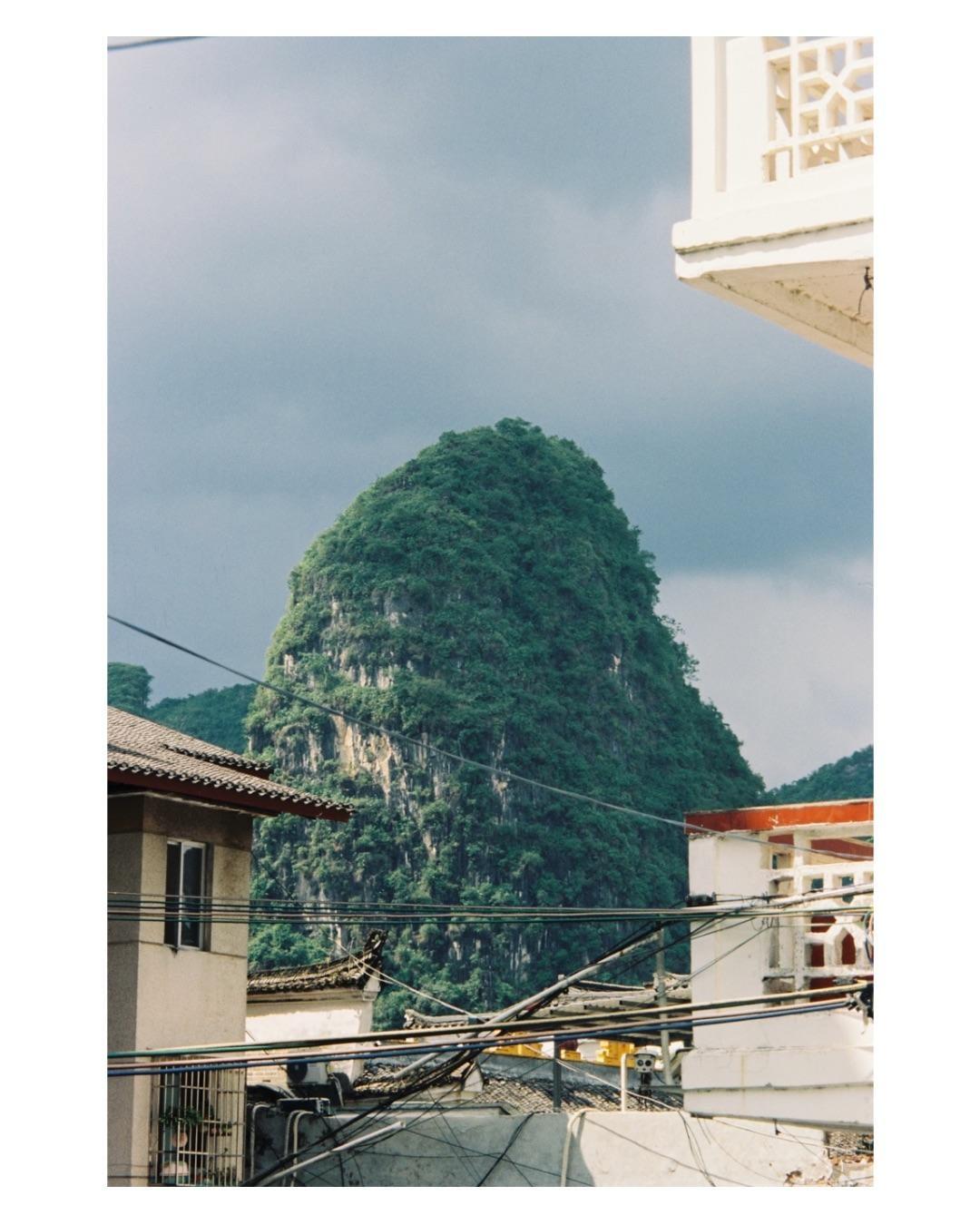 A lush green mountain rises behind buildings and a tangle of power lines under a cloudy sky.