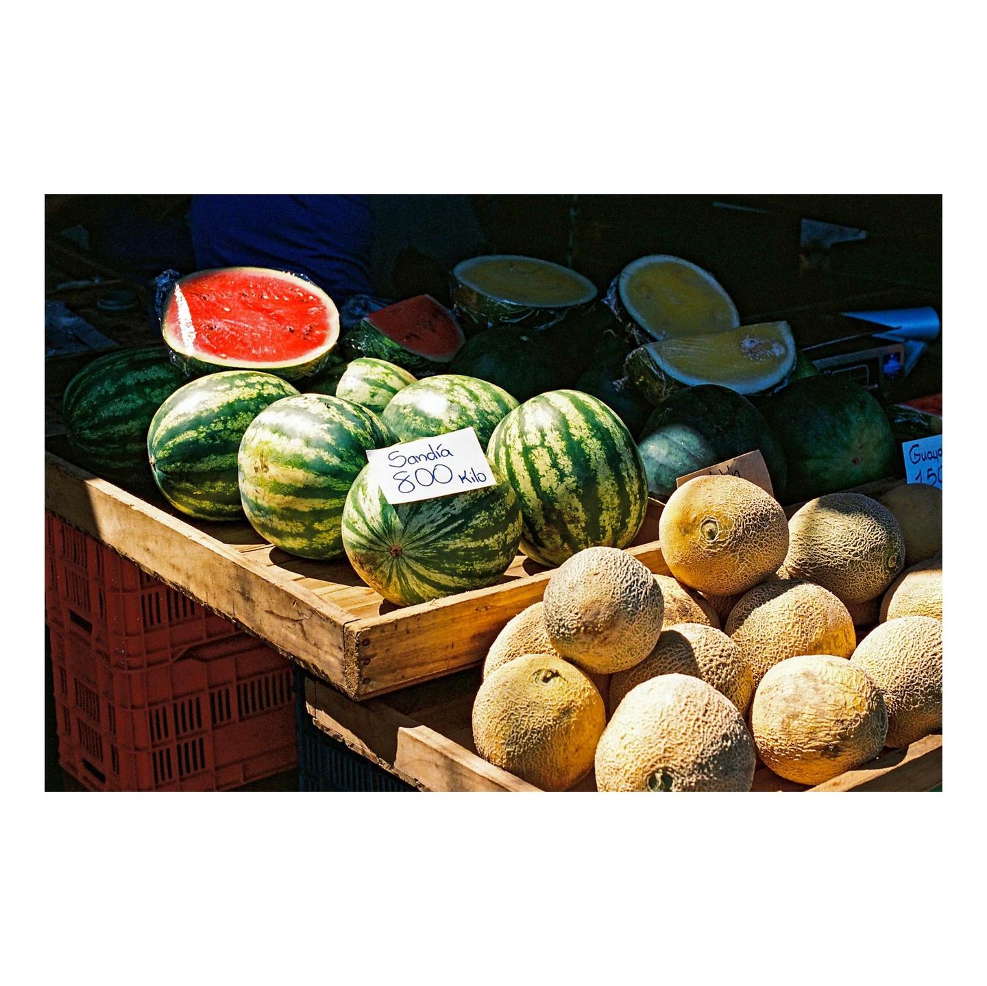 A market stall displays watermelons and cantaloupes in wooden crates.
