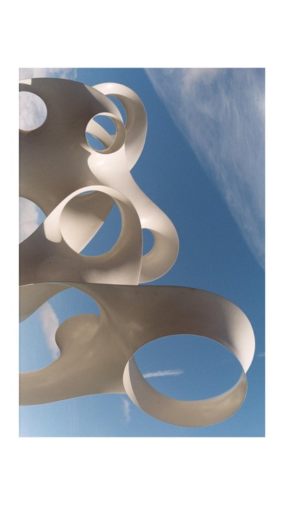 A white abstract sculpture with circular openings is set against a clear blue sky with wispy clouds.
