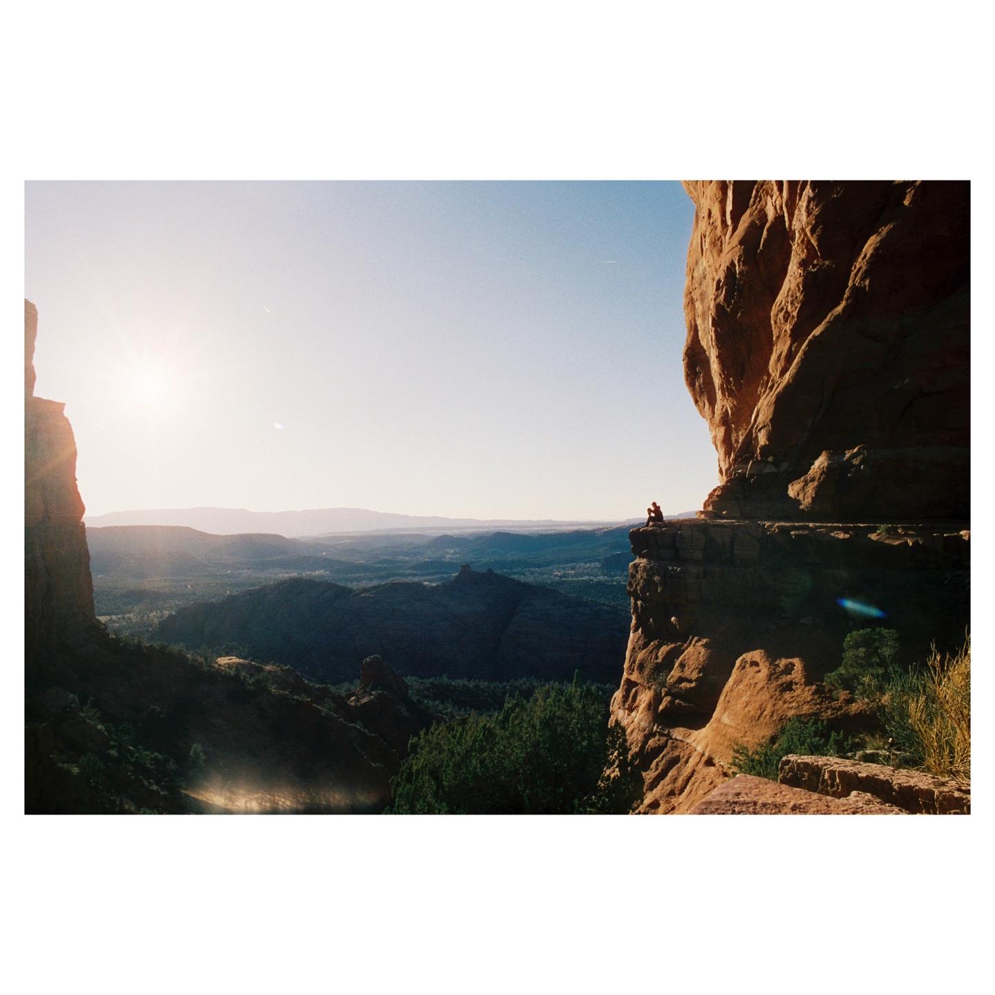 A person sits on a cliff overlooking a vast landscape with mountains and trees under a bright sky.