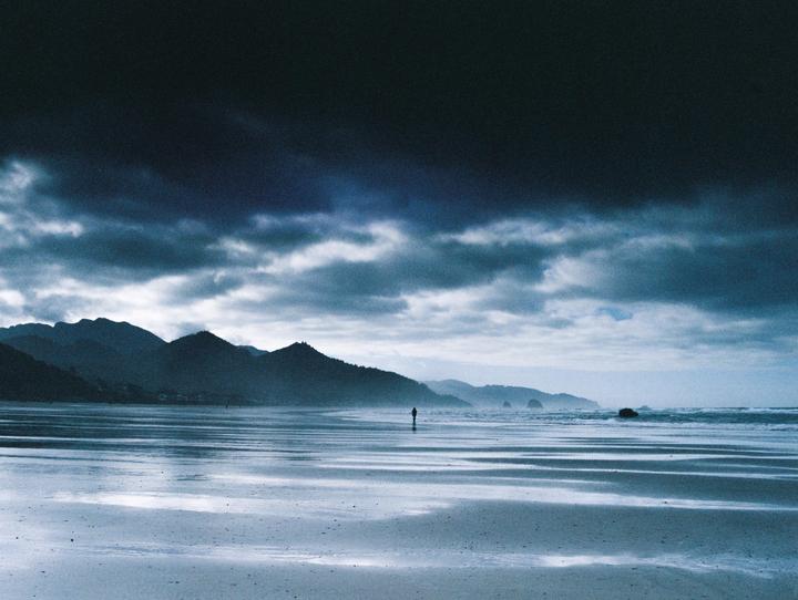 A lone figure stands on a wide, wet beach under a dramatic, cloudy sky with mountains in the distance.