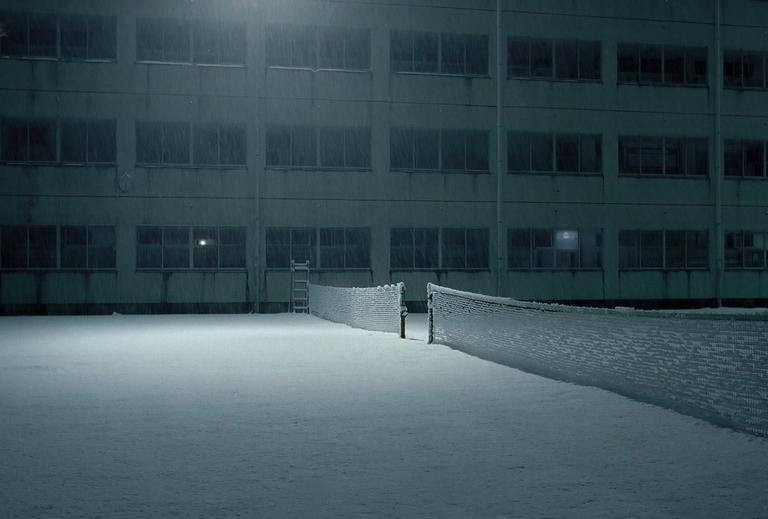 A snow-covered tennis court and building are illuminated by a single light at night.