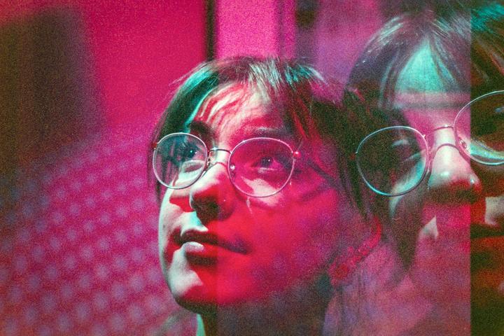 A woman with glasses looks up, illuminated by pink and blue light, with a faint double exposure of her face.