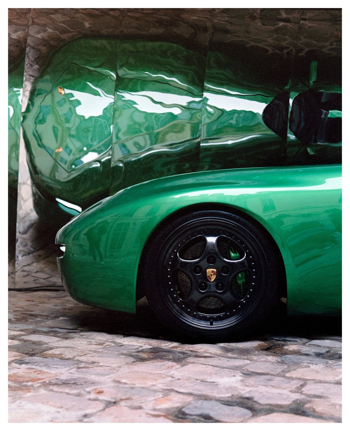 A green car is parked on a cobblestone surface next to a reflective green wall.