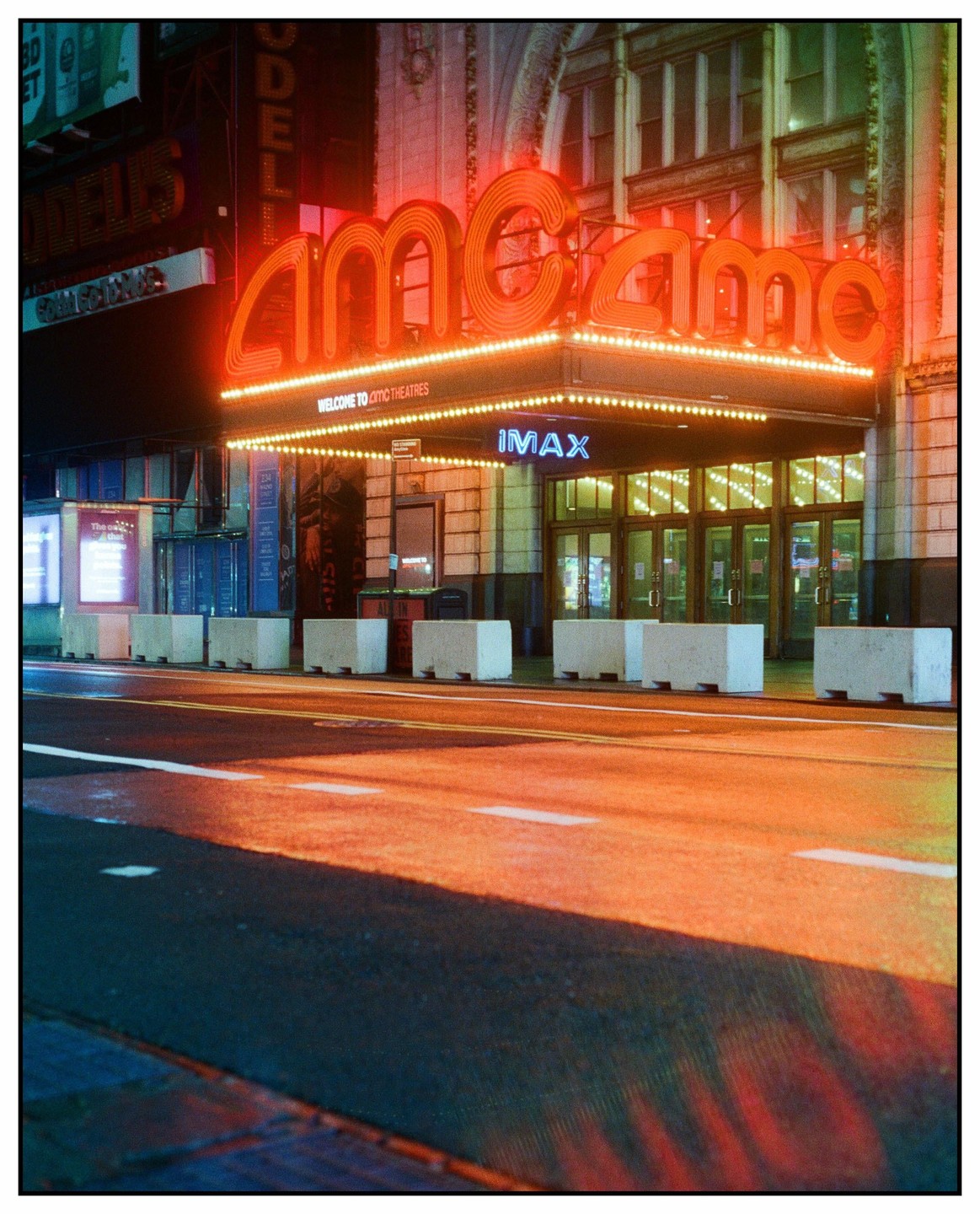 A movie theater with a large neon sign is illuminated at night, casting reflections on the wet street.