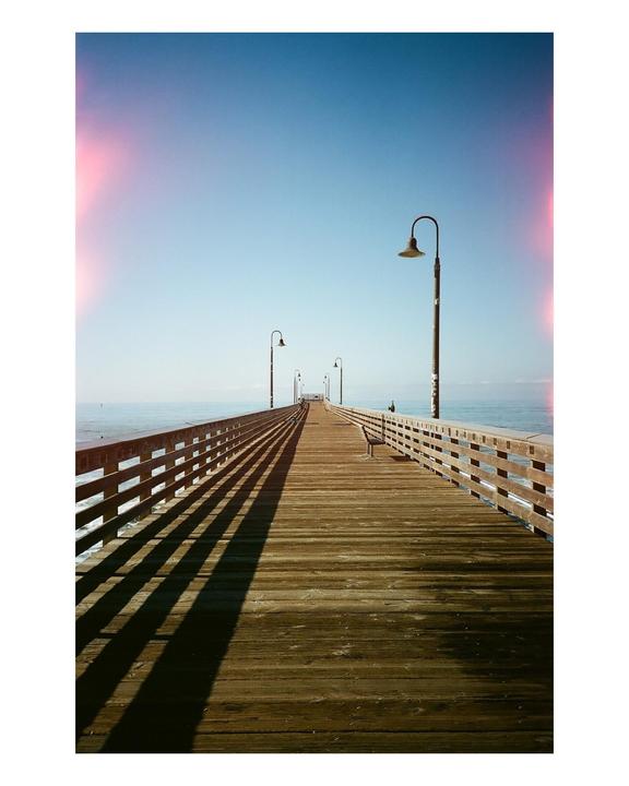 A long wooden pier stretches into the ocean under a clear blue sky, with shadows from the railing and lampposts extending across its surface.