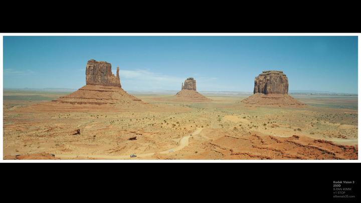 Three large rock formations stand in a vast desert landscape under a clear sky, with a dirt road winding through the foreground.