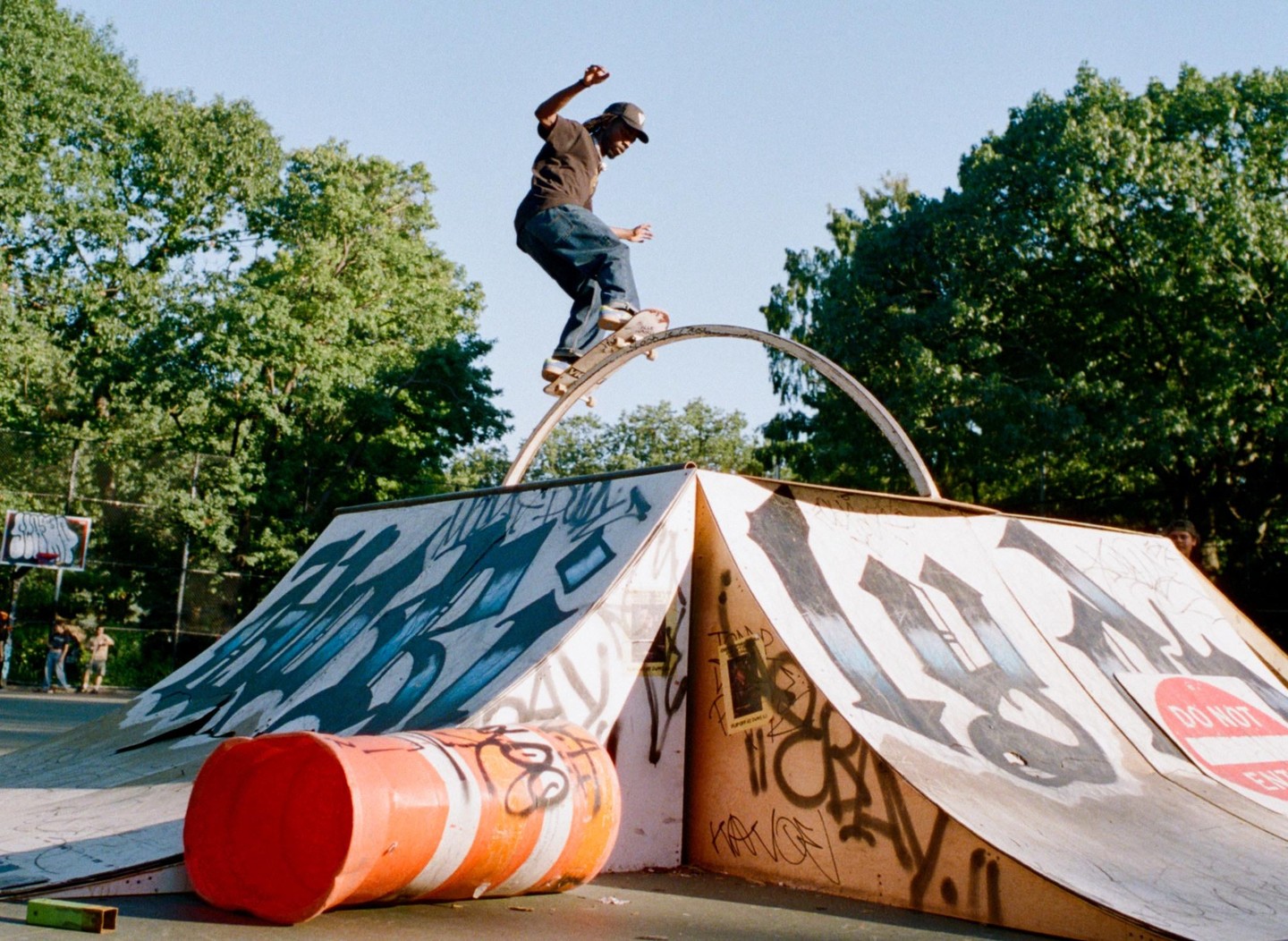A man on a skateboard balances on a metal arch at a skate park with graffiti-covered ramps and an orange barrel.