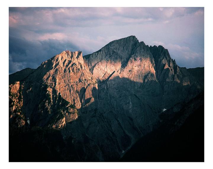 A mountain peak is illuminated by the setting sun under a cloudy sky.