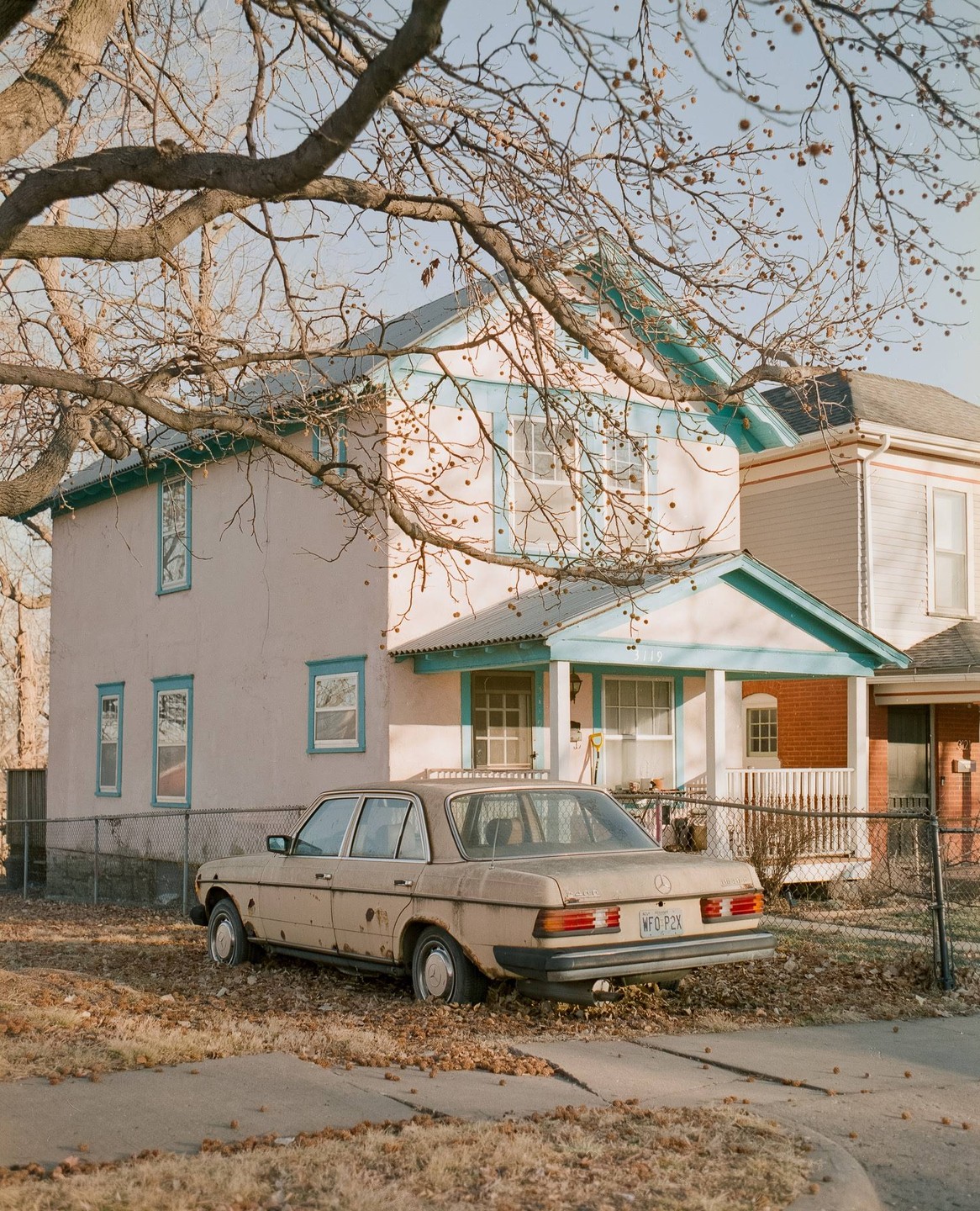 A beige car is parked in a yard with fallen leaves in front of a two-story house with blue trim.