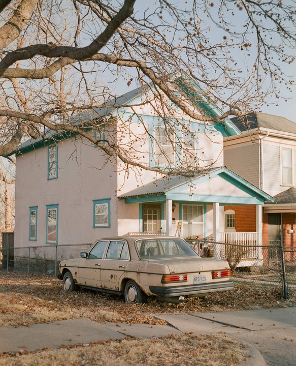 A beige car is parked in a yard with fallen leaves in front of a two-story house with blue trim.