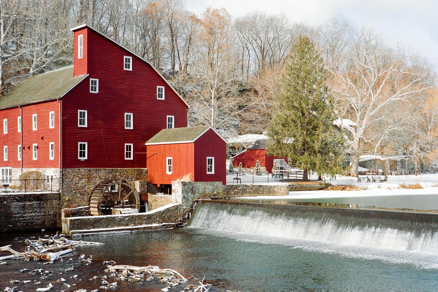 A red mill building with a water wheel stands next to a river with a waterfall, surrounded by snow-covered trees.