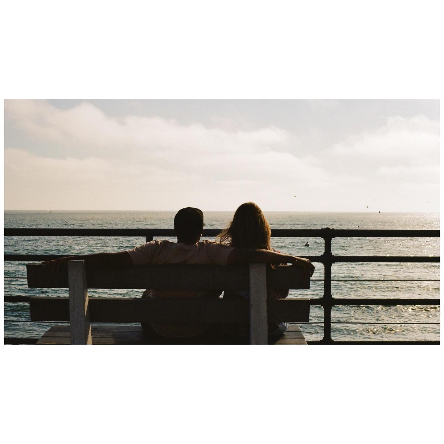 A couple sits on a bench overlooking the ocean from a pier.