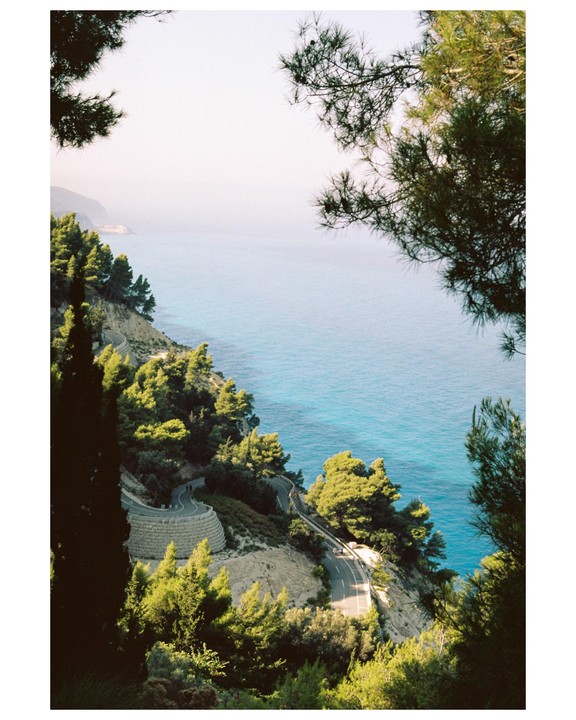 A winding road follows the coastline next to a turquoise sea, framed by pine trees.