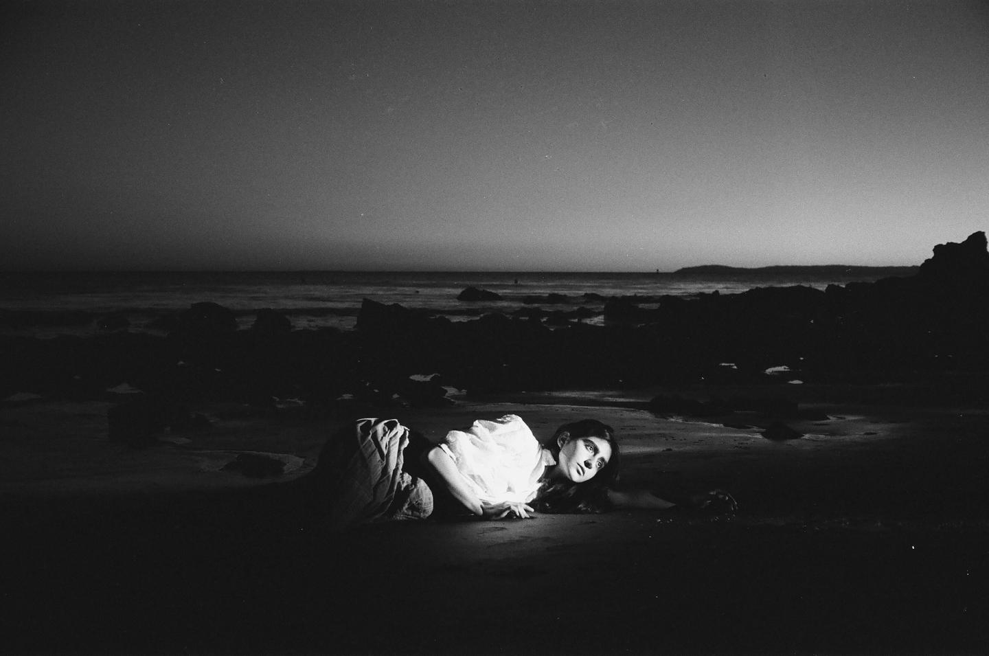 A woman lies on a dark beach with rocks and ocean in the background.